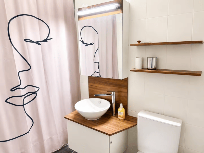 Modern bathroom with a white sink, wooden cabinet, mirror, and toilet. Features a sleek faucet, wooden shelves, and a decorative shower curtain.