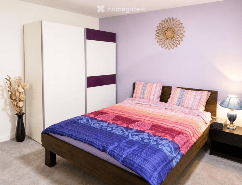Bedroom with a wooden bed, colorful bedding, nightstand with lamp, vase with dried flowers, and decorative wall art.