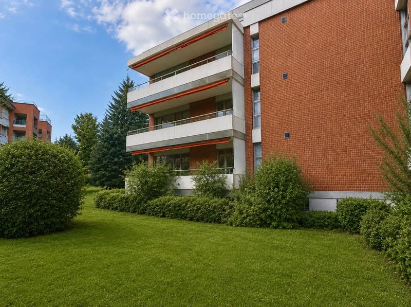 Modern multi-story apartment building with red brick facade, white balconies, and green lawn in front.