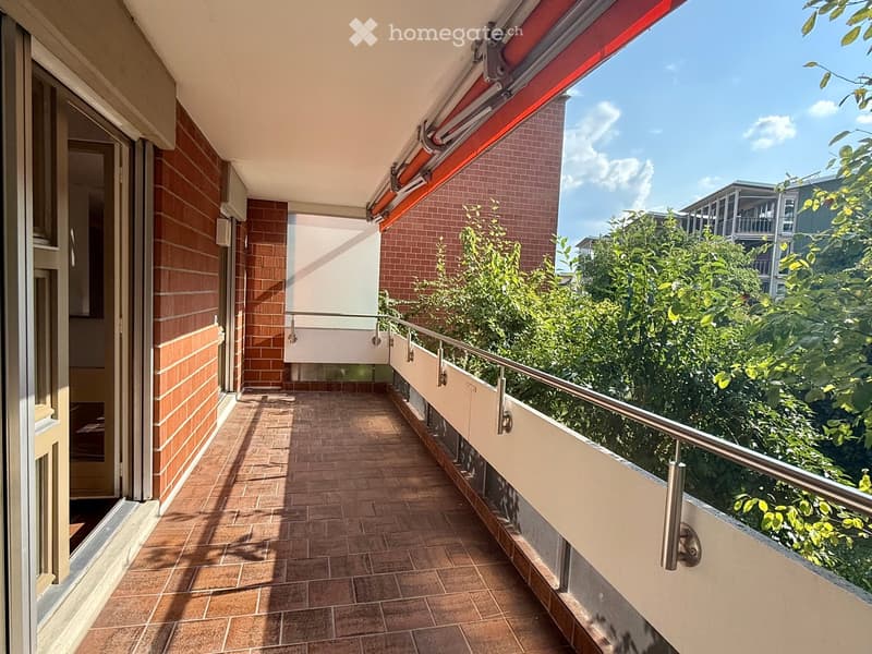 Balcony with tiled floor, metal railing, and red awning, overlooking greenery and neighboring buildings.