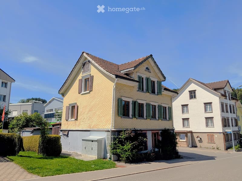 Two-story house with yellow siding, green shutters, and a tiled roof. The house has multiple windows and a small garden area in front.