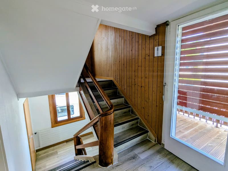 Wooden staircase with a curved handrail leads to an upper level, flanked by wooden panel walls and a large window with blinds. The floor is light-colored wood, and the ceiling is white with a sloped design.