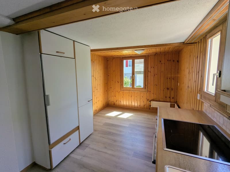 Modern kitchen with light wooden cabinets, a refrigerator, a stove, and a sink. The space features wooden walls and flooring with large windows allowing natural light.