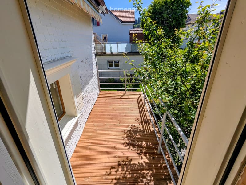 Wooden balcony with white railing, surrounded by greenery and neighboring buildings.