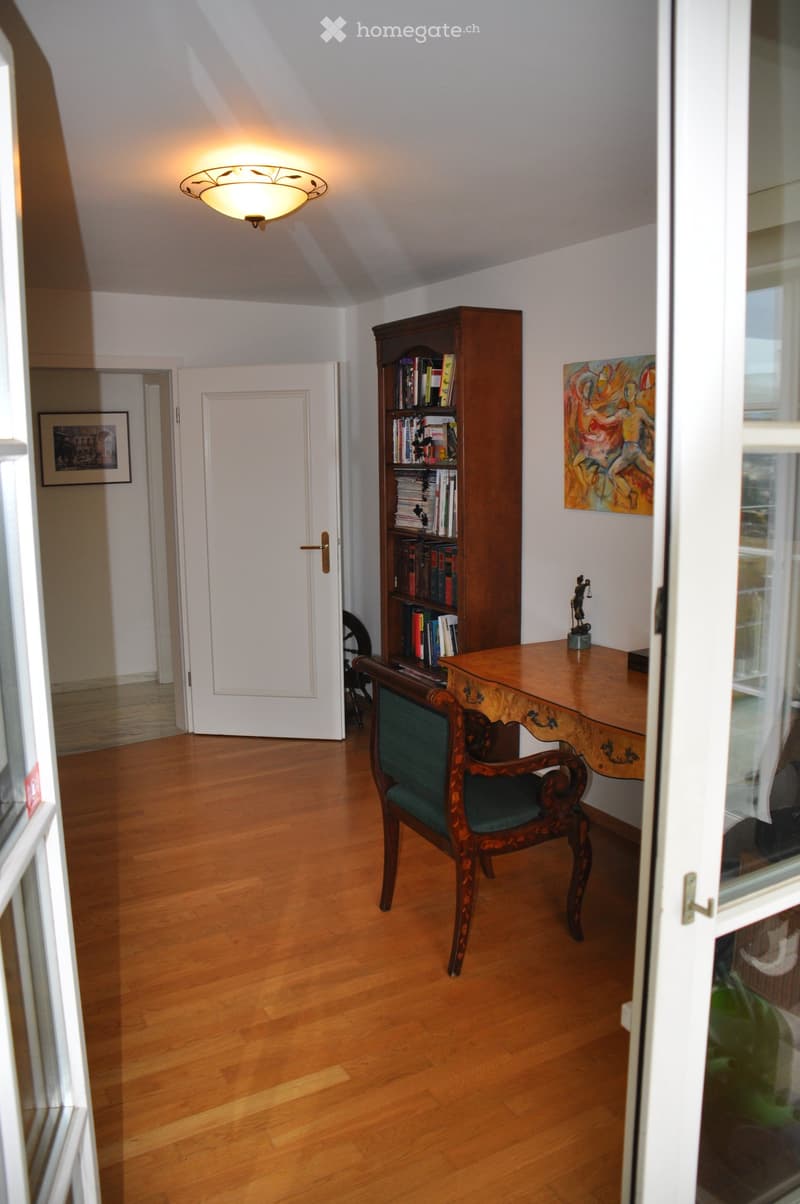 A well-lit room with wooden flooring, a wooden desk, a green upholstered chair, a tall wooden bookcase filled with books, and a painting on the wall.