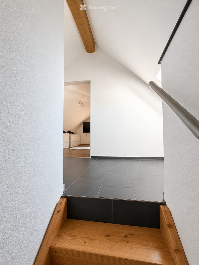 Modern staircase with wooden steps, metal railing, and grey tiled landing leading to an adjacent room with white walls and wooden flooring.