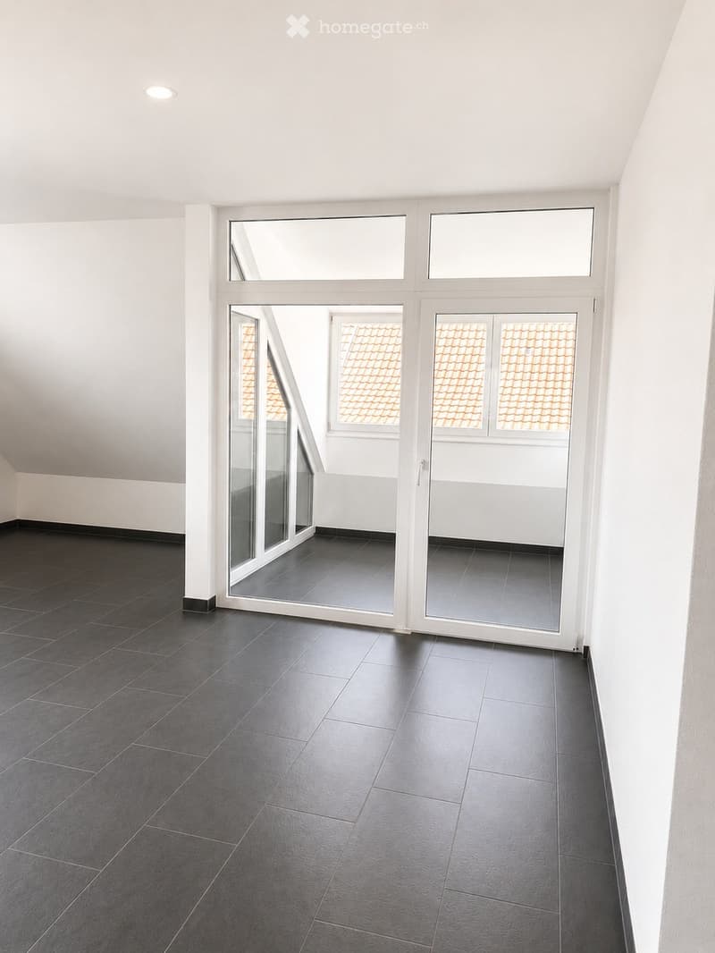 Empty room with dark grey tiled floor, white walls, and large windows with white frames, offering a view of a tiled roof outside.