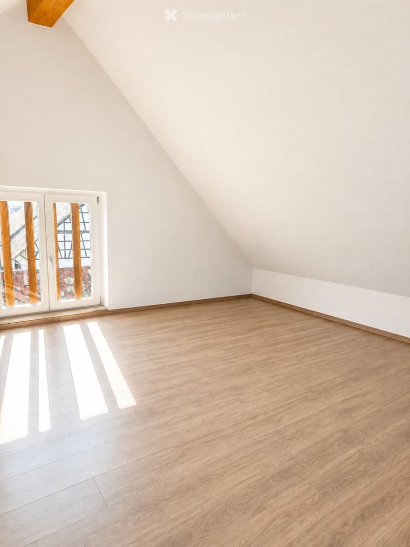 Empty attic room with wooden flooring, white walls, and large windows with wooden frames, allowing natural light to enter.