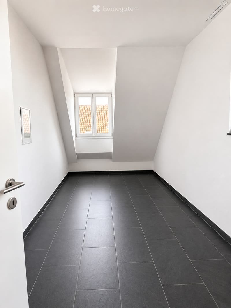 Empty room with dark grey tiled floor, white walls, and a window at the far end. The room has a door on the left side with a silver handle and a small picture frame on the wall.