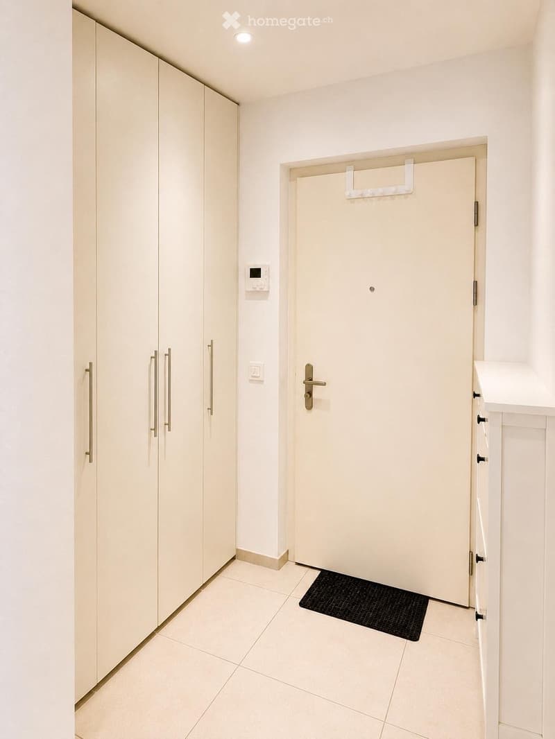 Clean, modern hallway with white walls, tiled floor, built-in wardrobes, and a closed door.
