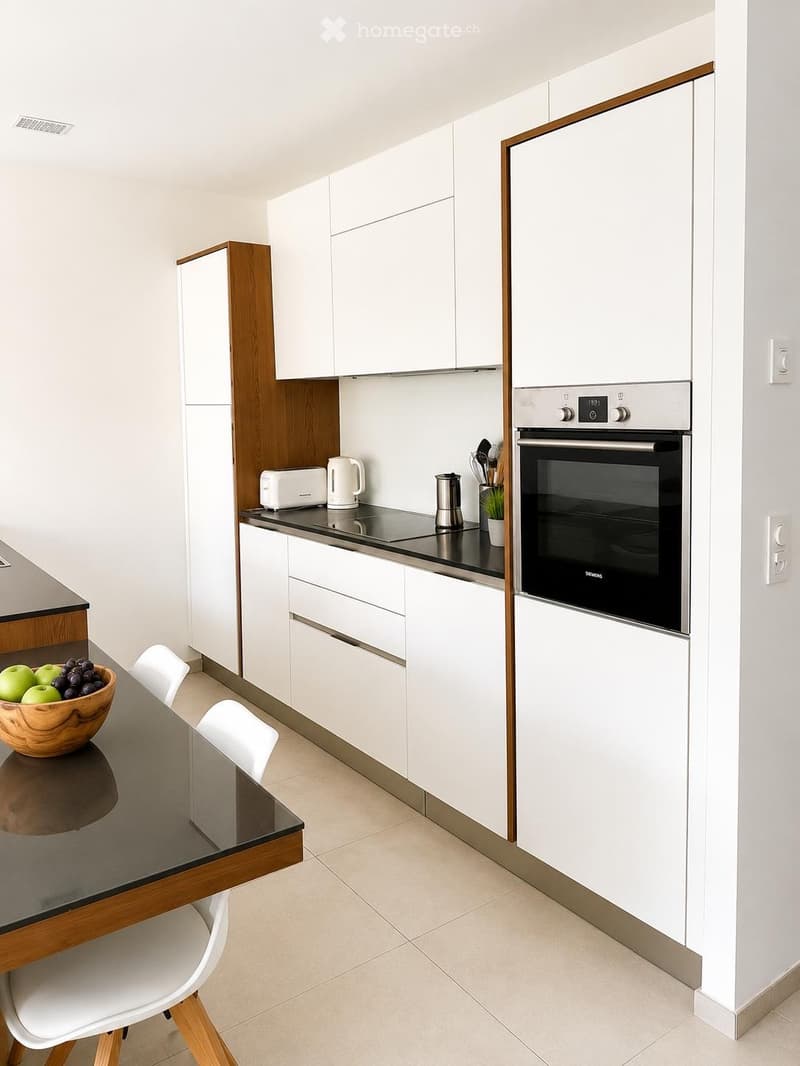 Modern kitchen with white cabinets, stainless steel appliances, a glass dining table with chairs, and a bowl of fruit on the counter.