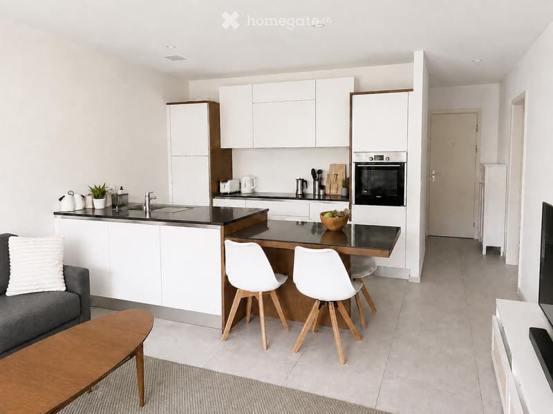 Modern kitchen and living area with white cabinets, black countertops, a grey sofa, a wooden coffee table, and a dining table with chairs.