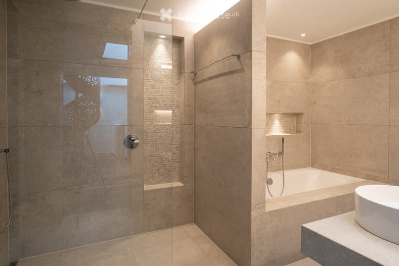 Large, modern bathroom with a glass-enclosed shower, a bathtub, and beige tile walls and floors. The bathroom has recessed lighting and a window providing natural light.