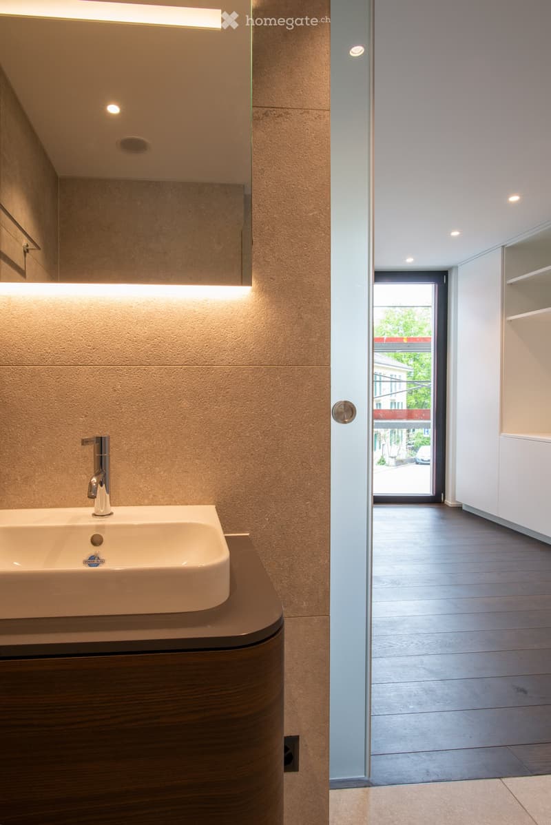 Spacious bathroom with beige stone tiles, a large vanity with a sink, and a window overlooking the exterior of the building.