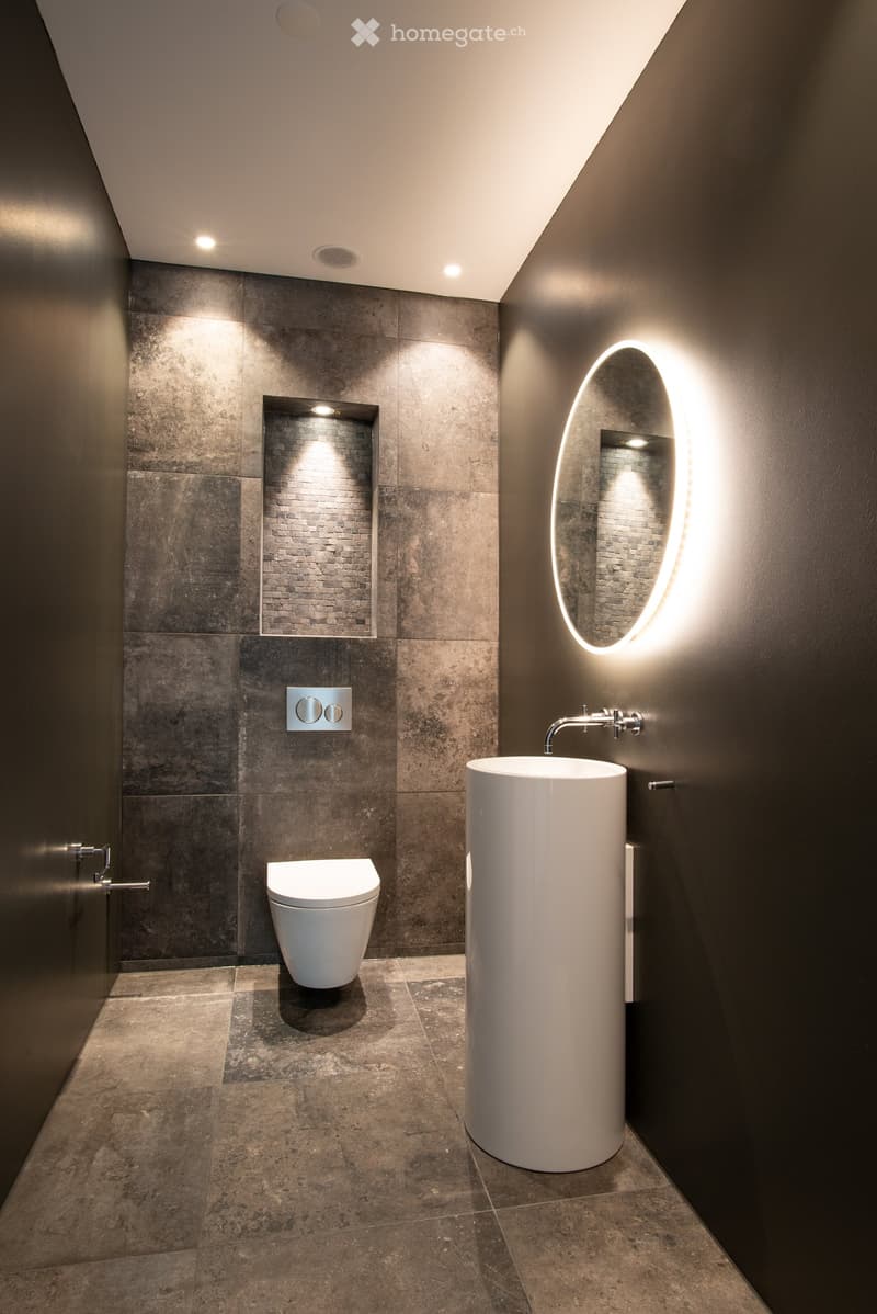 Modern bathroom with gray tile walls, a freestanding white sink, a toilet, and a large oval-shaped backlit mirror. The room has recessed lighting and a minimalist design.