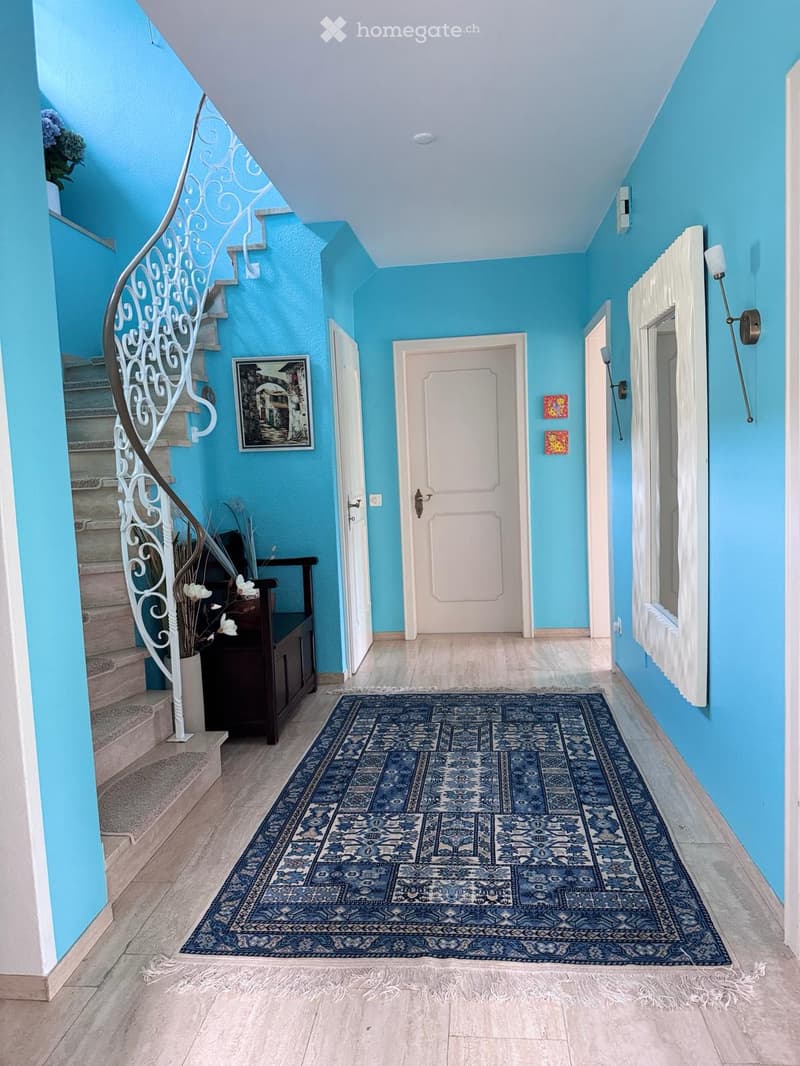 Bright hallway with blue walls, wooden floor, patterned rug, staircase with white wrought-iron railing, and a small bench with a vase of white flowers.