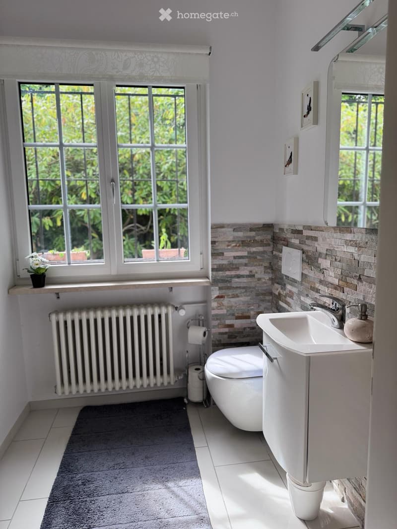 Modern bathroom with white tiles, a sink, toilet, radiator, and a large window with a view of greenery.