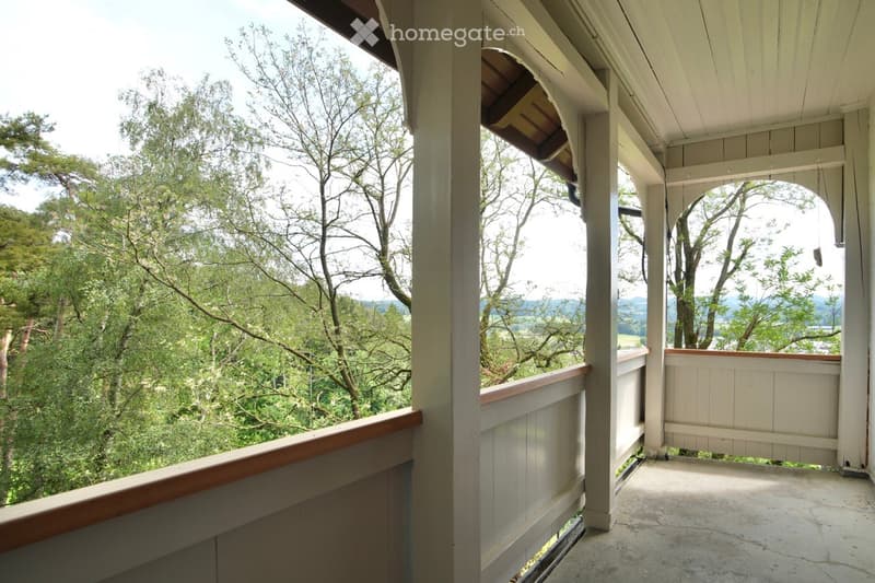 A spacious wooden balcony with white railings and a view of a lush, green forest and distant mountains.