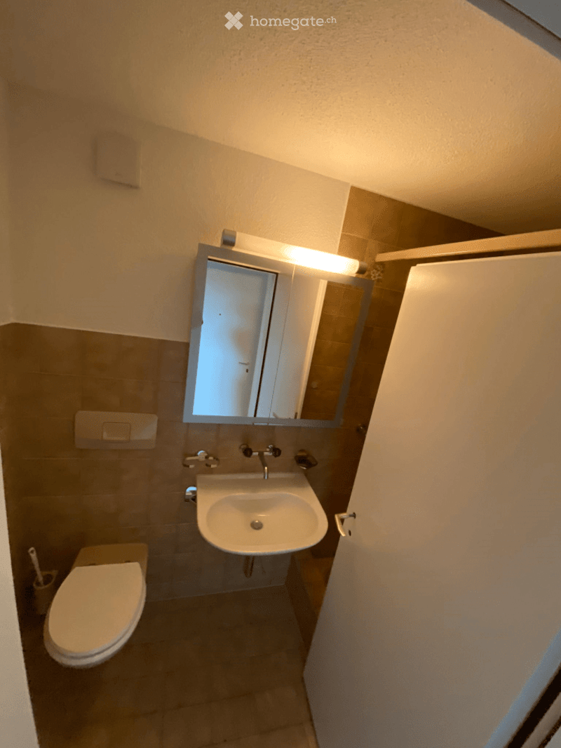 Modern bathroom with a toilet, sink, mirror, and faucet. The walls are tiled in neutral tones, and the space is compact with a clean, contemporary design.