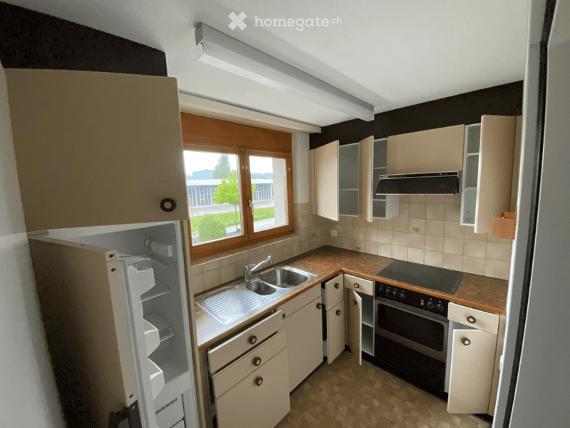 A small kitchen with light-colored cabinets, a sink with a faucet, a refrigerator, an oven, and a stove. The space has tiled walls and a window offering a view of the outdoors.