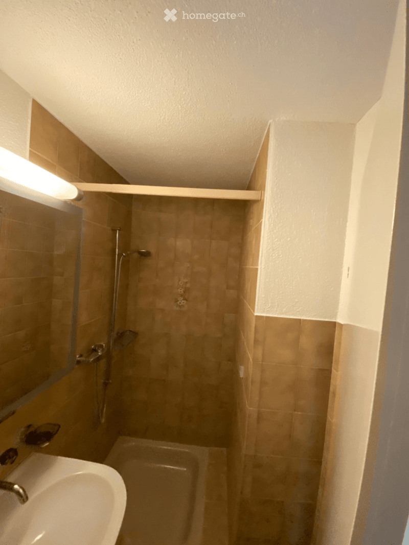 Small bathroom with beige tiles, a sink, a bathtub, a shower, and a mirror. The space is compact with a built-in shelf above the bathtub.