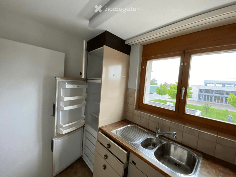 Modern kitchen with light-colored cabinets, a stainless steel sink, and an open refrigerator. The space features wooden countertops and large windows offering a view of the outdoors.