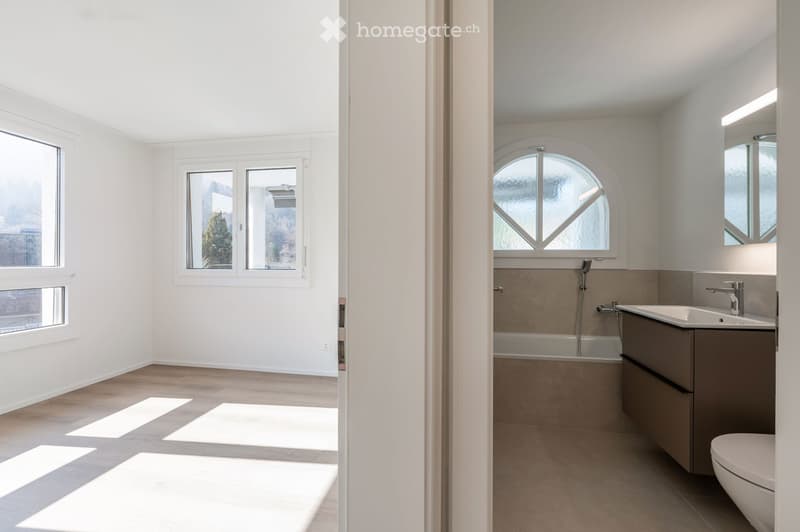 Modern bathroom with white walls, wooden floor, and large windows. Features a bathtub, sink, and toilet. The room is currently empty.