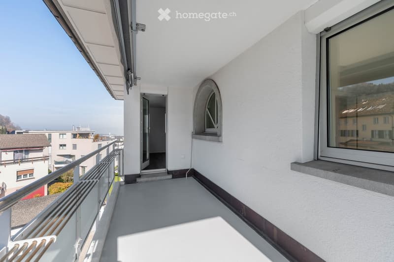 Empty balcony with glass railing, white walls, and a window with a view of the surroundings.