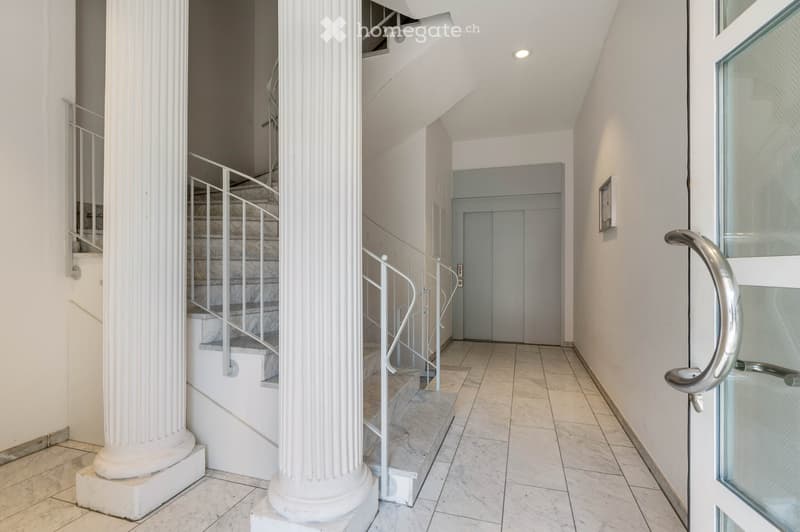 A corridor with marble flooring, white walls, and two large white columns. A staircase with marble steps and metal handrails leads to the upper floor. A door with a glass panel and a silver handle is on the right side.