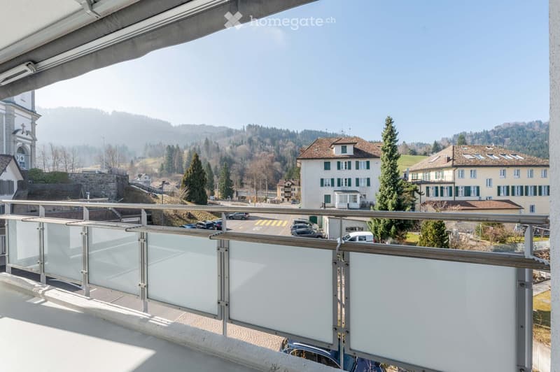 Balcony with a metal railing, overlooking a street with parked cars and buildings.