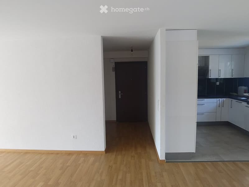 Empty hallway with wooden flooring, white walls, and a door leading to another room. A kitchen area is visible at the end of the hallway with white cabinets and a countertop.