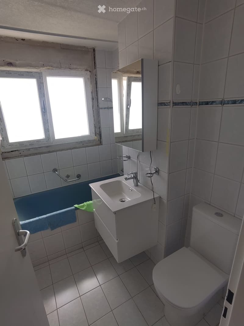 Bathroom with tiled walls and floor, a white sink with a chrome faucet, a toilet, a mirror, and a small cabinet. Blue towels are draped over the edge of the bathtub.
