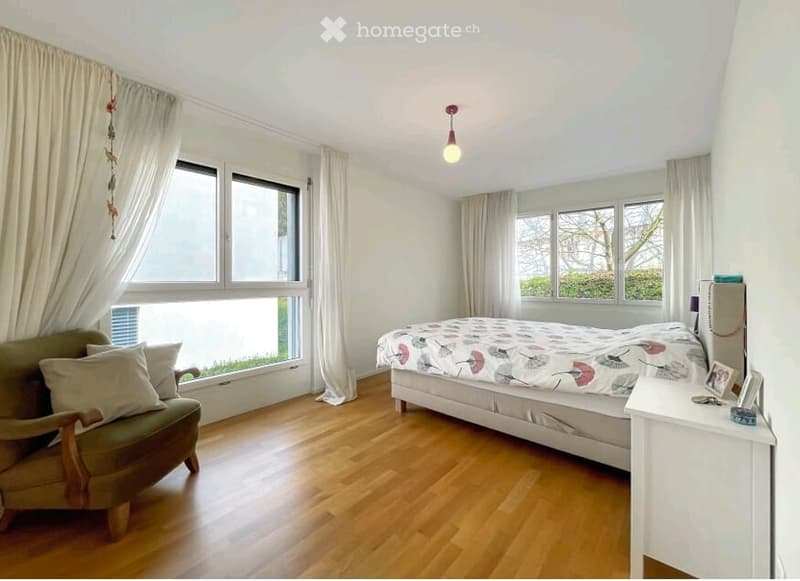 Bright bedroom with wooden flooring, large windows with curtains, a double bed with a patterned duvet, a green armchair, and a nightstand with a lamp.