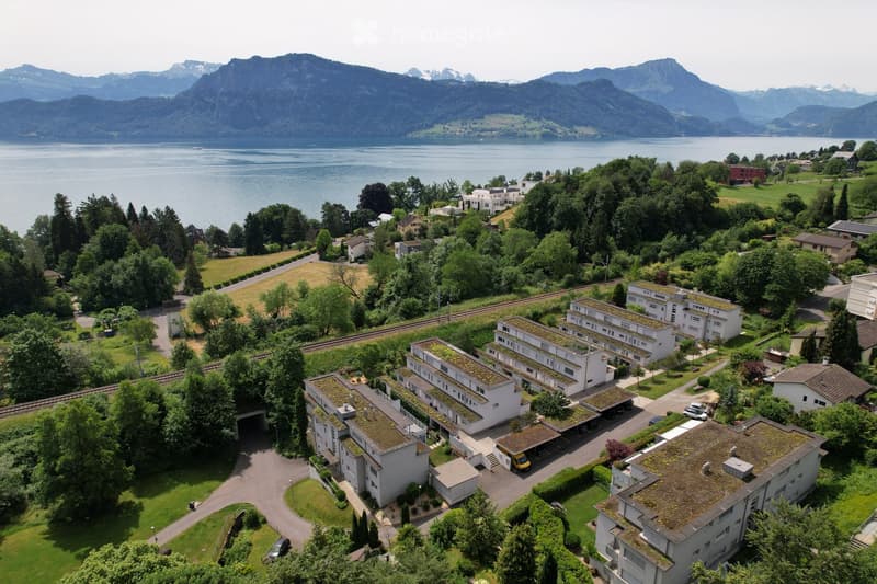 Aerial view of a residential complex with multiple buildings, surrounded by greenery, a parking area, and a scenic lake and mountain backdrop.