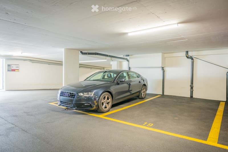 Underground parking space with a single car parked inside, marked parking spaces, and visible structural elements like pipes and lighting.