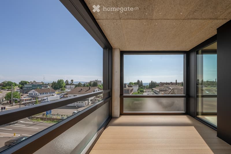 Modern balcony with wooden decking, large windows, and a view of a town with buildings, trees, and a road.
