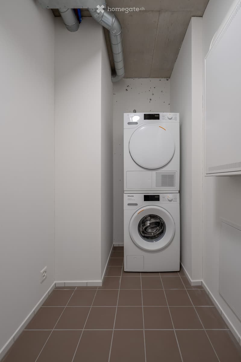 A small, narrow room with white walls and brown tiled flooring, featuring a stacked washing machine and dryer unit.