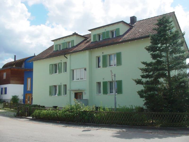 Two-story house with light green siding, green shutters, and a red-tiled roof. The house features multiple windows and a small garden area in front.