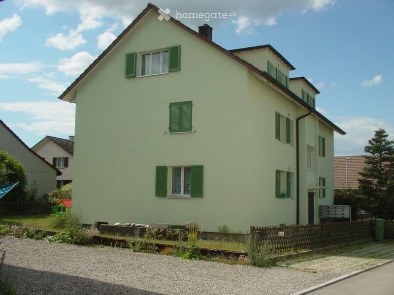 Two-story house with light yellow walls, green shutters, and multiple windows. The house has a pitched roof and is surrounded by a small garden and gravel driveway.