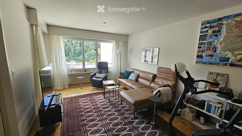 Modern living room with large windows, leather sofa, ottoman, armchair, coffee table, side tables, floor lamp, TV, bookshelf, and exercise bike.