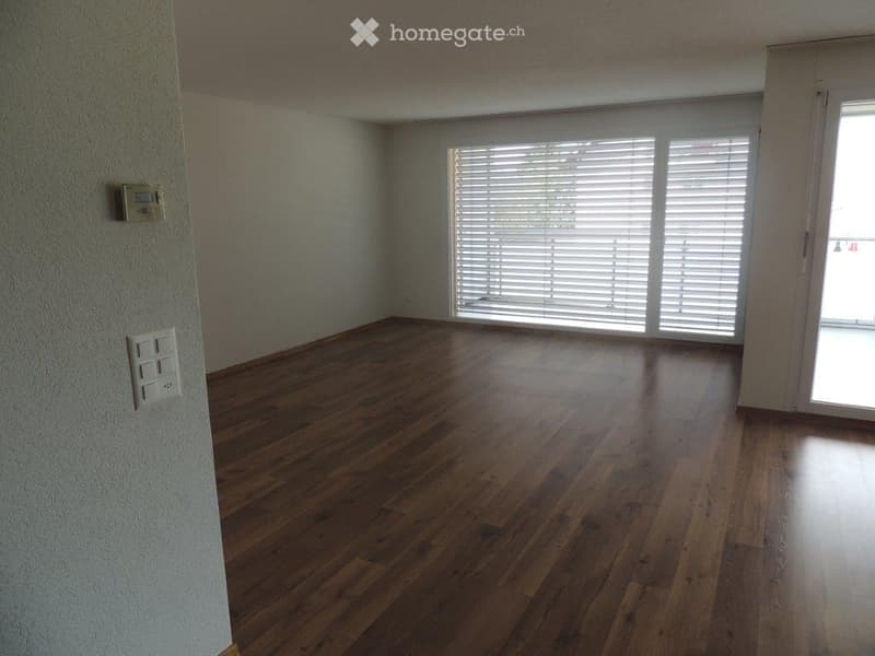 Empty room with wooden flooring, large windows with blinds, and a sliding glass door leading outside.