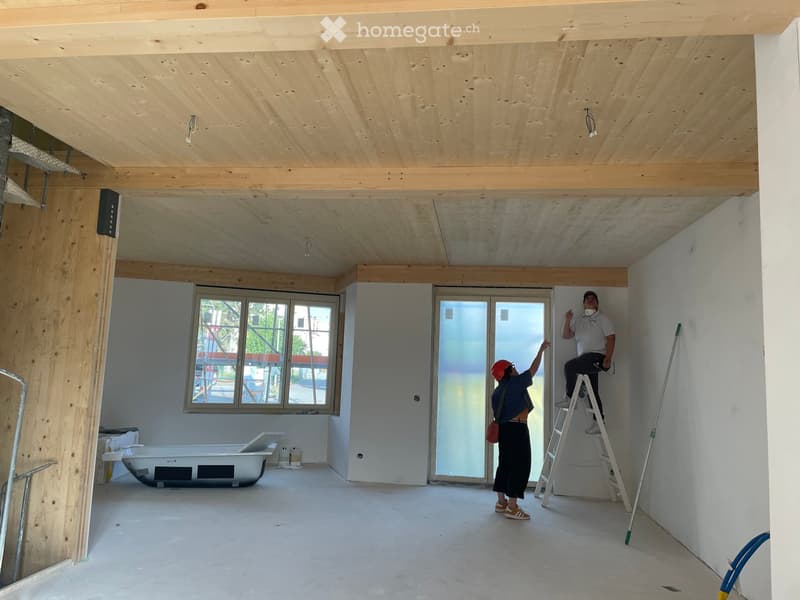 Empty room with exposed wooden beams, white walls, concrete floor, large windows, and a bathtub. Two workers are present, one on a ladder and the other pointing.