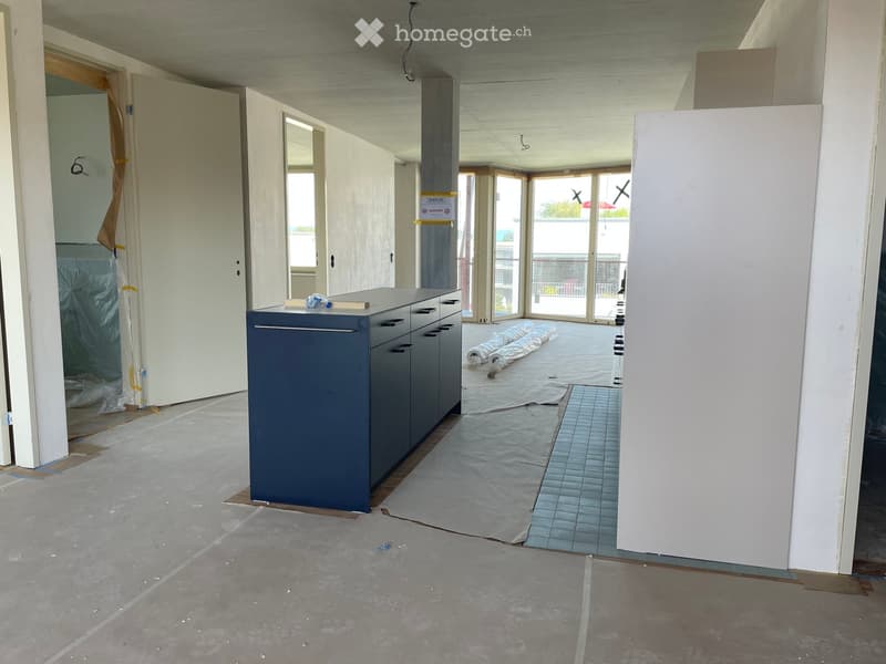 Empty room with unfinished concrete floor, exposed ceiling beams, and a partially installed kitchen cabinet unit. The space includes a doorway leading to another room and a large window with a view of a balcony area.