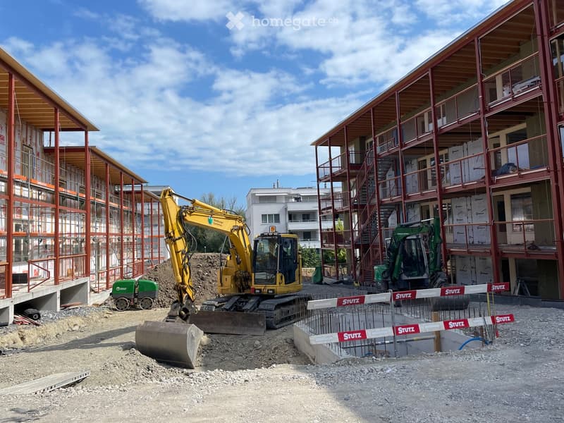 Construction site with two multi-story buildings under construction, featuring exposed wooden beams and red metal framing. Heavy machinery, including excavators, is present, along with construction barriers and scaffolding.