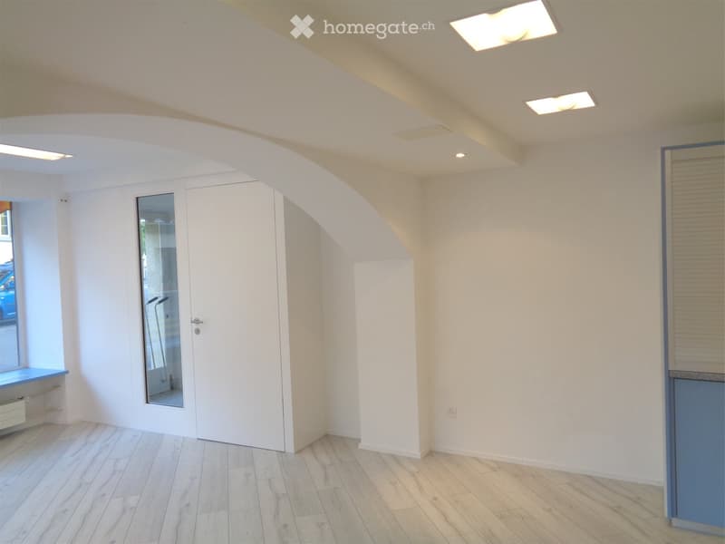 Empty room with light-colored wooden flooring, white walls, and a large window with blinds. The ceiling has recessed lighting, and there is a white door with a glass panel leading to another space.