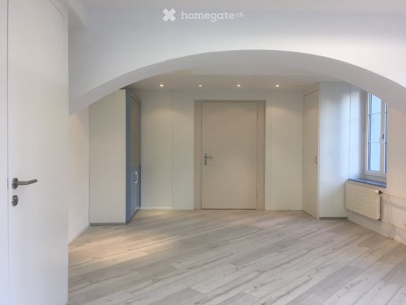 Empty room with light wooden flooring, white walls, and several doors leading to other spaces.