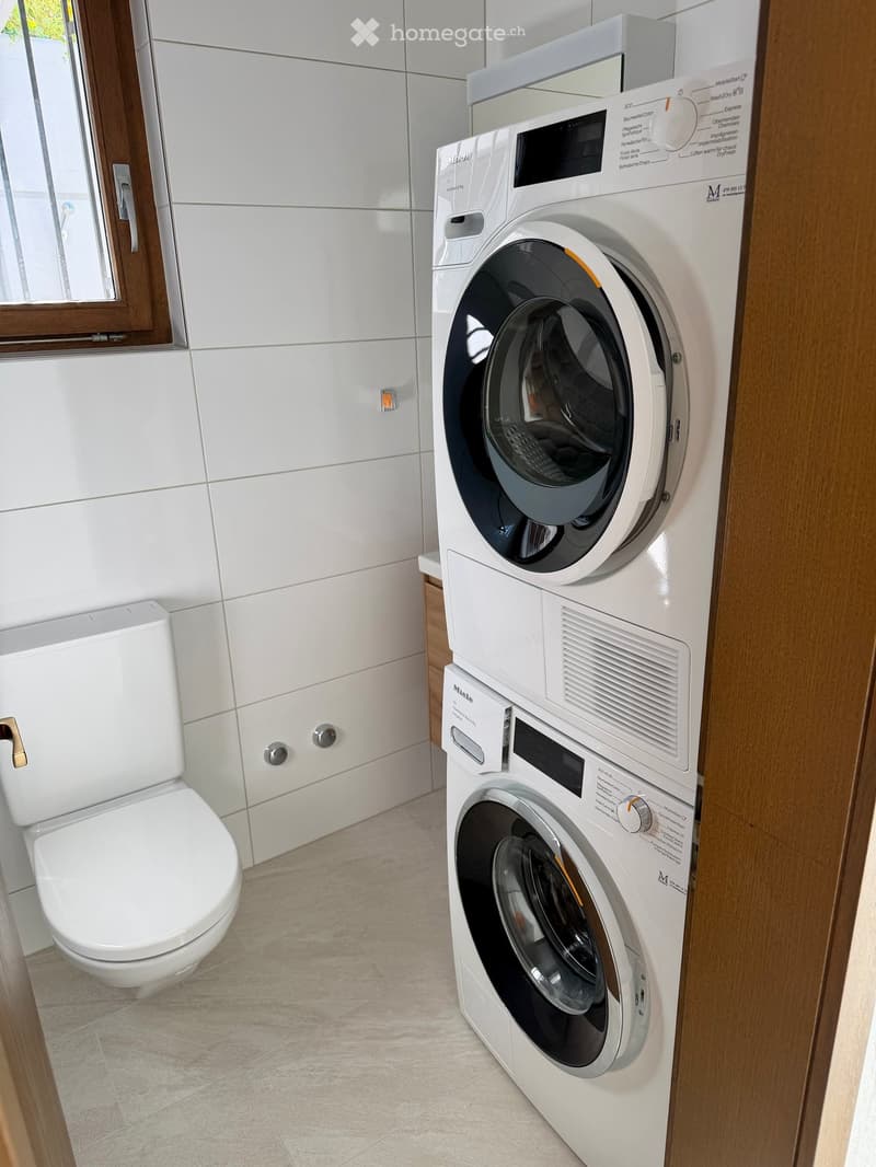 Modern bathroom with a toilet, washing machine, and dryer, white tiled walls, and a window with wooden frame.