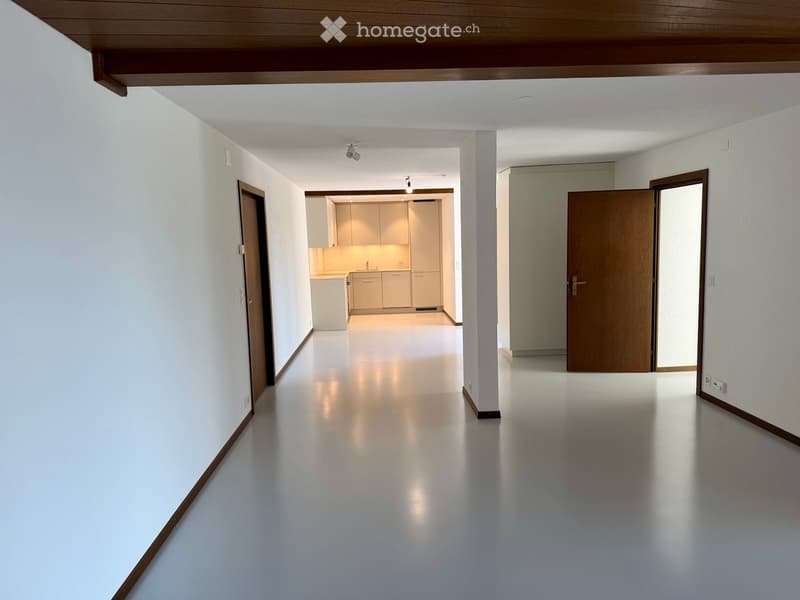 Empty hallway with white walls, polished floor, and doors leading to rooms, including a kitchen area with cabinets and a countertop.