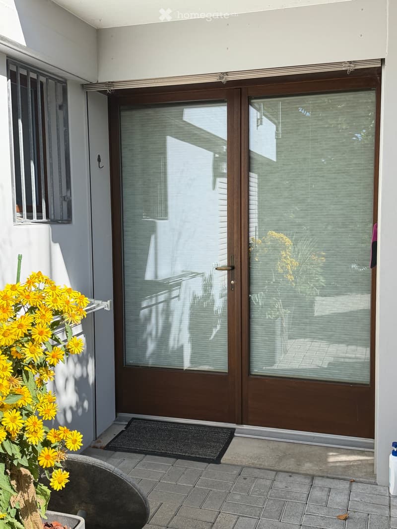 Balcony with a wooden sliding door, a doormat, and a potted plant with yellow flowers.