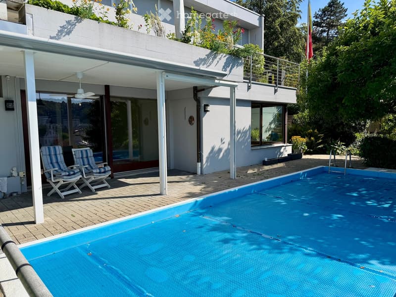 Outdoor area with a swimming pool, two striped lounge chairs, and a modern house with a balcony and greenery.